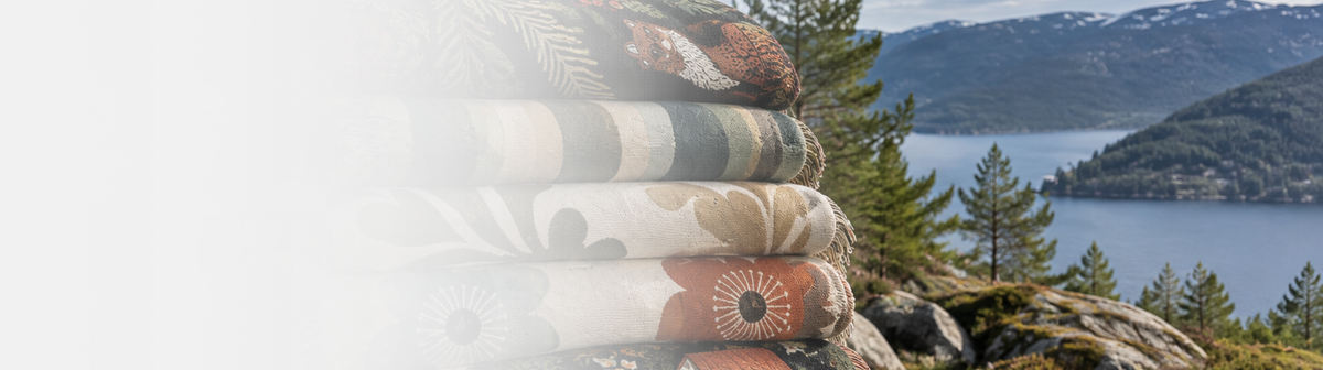 A stack of folded patterned textiles or blankets with floral and geometric designs in beige, brown, and muted green tones, set against a scenic outdoor background of mountains, water, and trees. Havelli Nordic