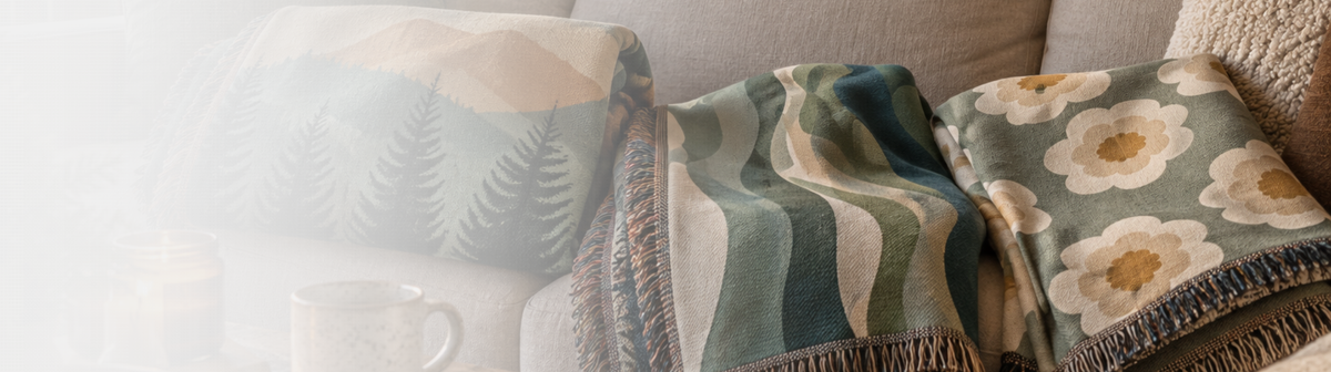 Three cozy woven throw blankets draped over a light-colored sofa. The left blanket has a muted green fern pattern on a cream background, the middle blanket features wavy stripes in teal, sage green, and cream, and the right blanket displays large cream flowers with yellow centers on a dusty green base. Havelli Nordic