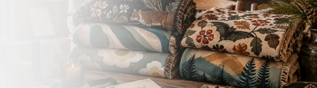 A stack of woven tapestries and blankets with rustic floral and landscape patterns in muted earth tones, set against a softly lit, blurred interior background. Gift havelli nordic